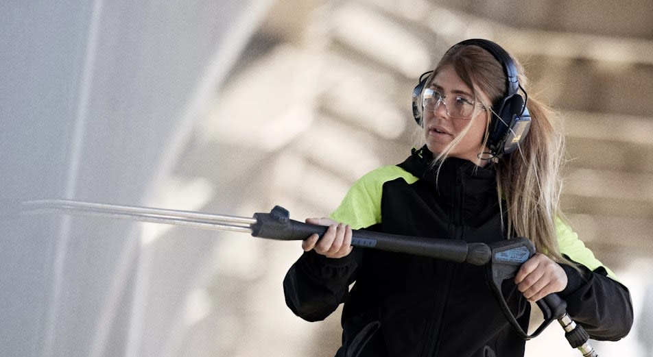 En kvinna i arbetskläder, skyddsglasögon och Helllberg-hörselskydden Xstream MP högtryckstvättar en vägg.