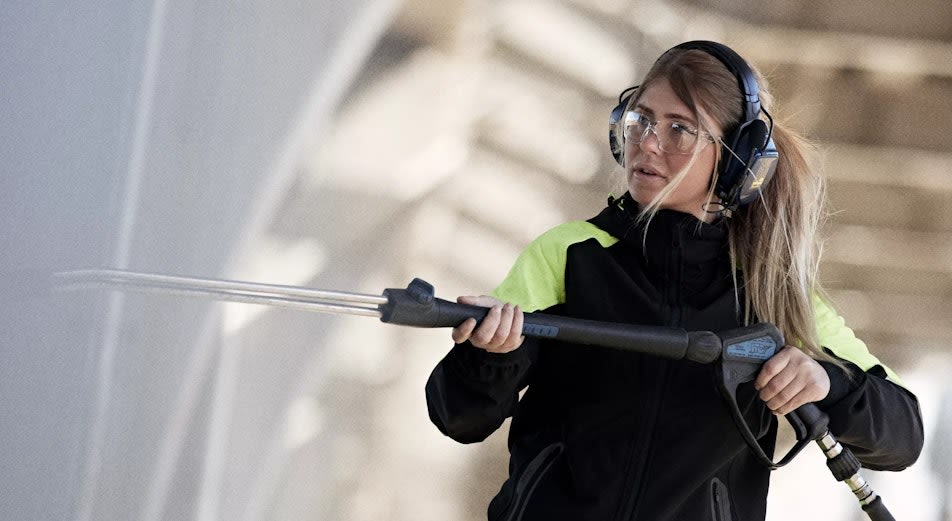 Nainen työvaatteissa, suojalaseissa ja Hellbergin Xstream MP -kuulonsuojaimissa painepesee seinää.