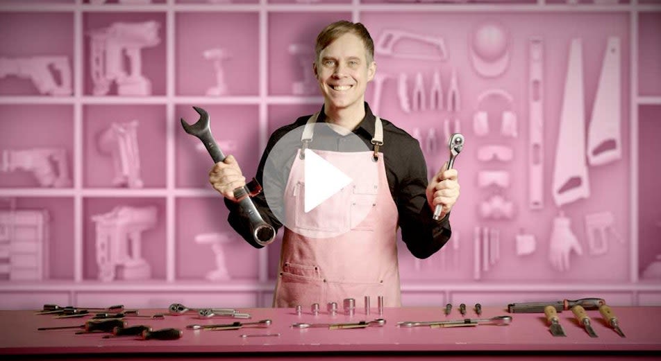 En man står och håller upp en stor uringnyckel och ett spärrskaft vid en desk med massor av verktyg.